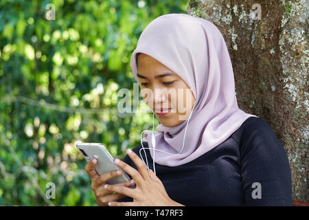 Close-up shot della donna musulmana in hijab utilizza lo smartphone con auricolari. Le riprese del Malaysian giovane donna o studente guardando sullo smartphone. Foto Stock