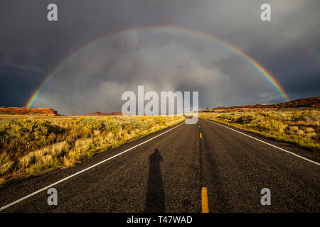 L'ombra del fotografo mostra sulla strada sotto un arcobaleno pieno e thuderstorm nubi su Scenic Highway 211 nel Parco Nazionale di Canyonlands, Sou Foto Stock