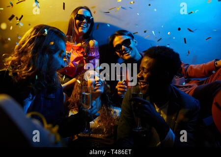 Gruppo di giovani amici multietnici eccitati seduti insieme sotto confetti e bevendo alcol mentre ridono e parlano a festa Foto Stock