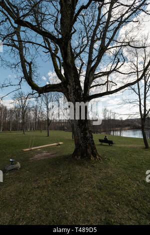 Lonely donna seduta su una panchina in un parco a pensare la vita guardando l'orizzonte -Liepupes Muiza manor in bella e soleggiata primavera meteo con blue Foto Stock