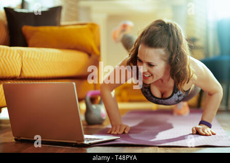 Sorridente donna sana in abiti di fitness nella moderna sala fitness guardare tutorial su internet tramite computer portatile e facendo pushups sul tappetino fitness. Foto Stock