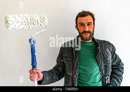 Giovani caucasici dai capelli marrone uomo con la barba e colorate in blu salopette da pantaloni di lavoro in piedi e tenendo la sua vernice tessuto Stick prolunga rullo P Foto Stock