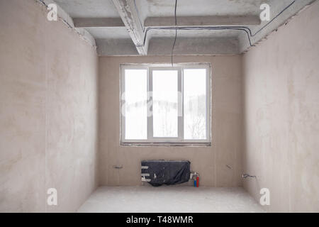 Stanza vuota con una finestra e una batteria in una casa in costruzione, pareti intonacate, un massetto sul pavimento, calcestruzzo multi-livello di lastre. Concetto di r Foto Stock
