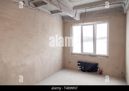 Stanza vuota con una finestra e una batteria in una casa in costruzione, pareti intonacate, un massetto sul pavimento, calcestruzzo multi-livello di lastre. Concetto di r Foto Stock