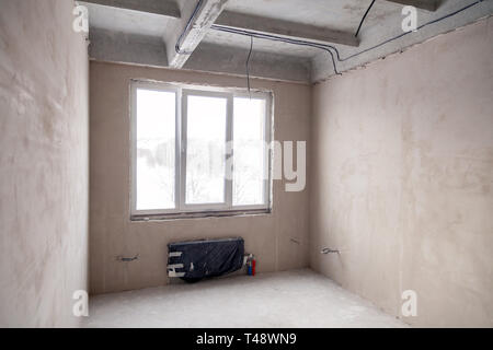 Stanza vuota con una finestra e una batteria in una casa in costruzione, pareti intonacate, un massetto sul pavimento, calcestruzzo multi-livello di lastre. Concetto di r Foto Stock
