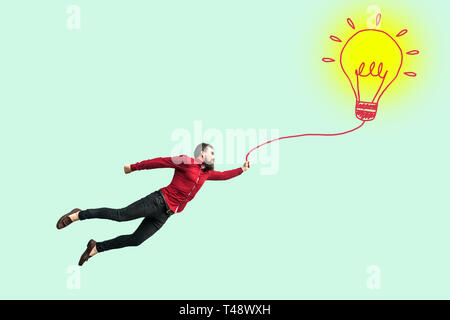 Giovane imprenditore di successo di volare con la sua perfetta idea. divertente successo giovane uomo barbuto in maglietta rossa azienda disegnò la lampadina lampada e battenti. interna Foto Stock