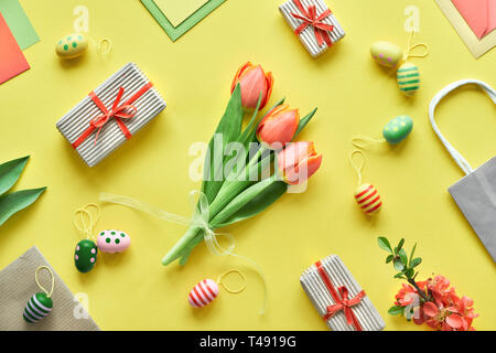 Pasqua laici piatta sulla carta gialla. Mazzo di tulipani, confezioni regalo, uova decorative e sacchi di carta, geometrica disposizione diagonale. Foto Stock