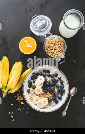 Sana colazione cereali con latte, mirtilli, banana su un calcestruzzo nero, sfondo tabella vista superiore piana e composizione dei laici Foto Stock