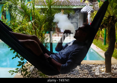 Uomo appoggiato in una amaca e vaping e scaricarsi da una sigaretta elettronica Foto Stock