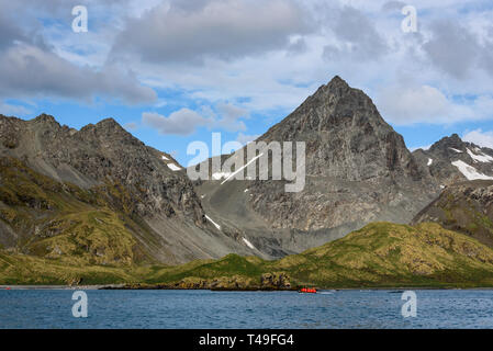 Bella giornata di sole a Coopers Bay con il grigio picco di montagna contro un bianco e blu cielo nuvoloso, gommone con i turisti in rosso cappotti Foto Stock