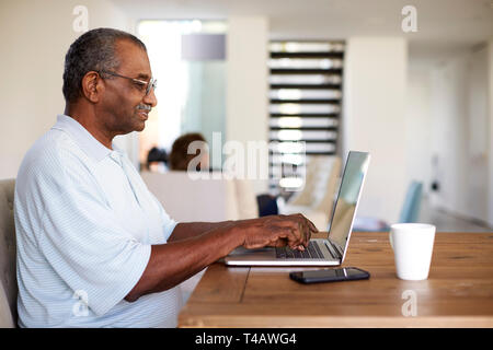 Senior uomo nero seduti a tavola utilizzando un computer portatile a casa, vista laterale Foto Stock