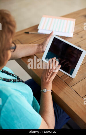 Senior donna seduta al tavolo utilizzando computer tablet, elevate sulla spalla, verticale Foto Stock