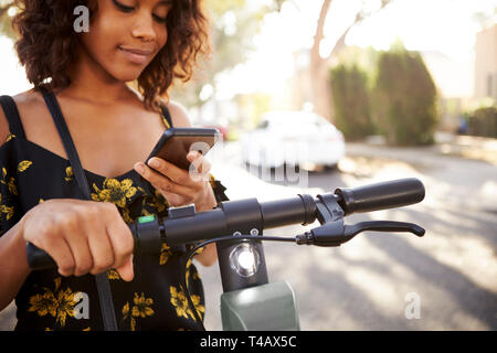 La millenaria nero donna in piedi su un scooter elettrico utilizza lo smartphone,close up
