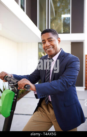Elegantemente vestito di mezza età uomo nero in piedi su scooter elettrico davanti a casa sua, close up Foto Stock