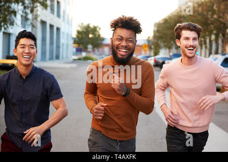 Anca tre giovani maschi adulti amici in esecuzione per divertirsi in una strada della città, vista frontale, close up Foto Stock