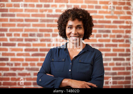 Ritratto di sorridente imprenditrice matura in piedi contro un muro di mattoni in ufficio moderno Foto Stock