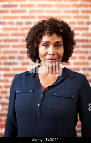 Ritratto di sorridente imprenditrice matura in piedi contro un muro di mattoni in ufficio moderno Foto Stock