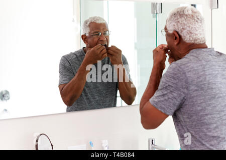 Senior uomo filo interdentale denti guardando la riflessione nello specchio del bagno indossando pigiami Foto Stock