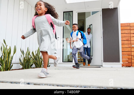 Emozionato i bambini in esecuzione fuori della porta anteriore sulla strada per la scuola guardato da padre Foto Stock