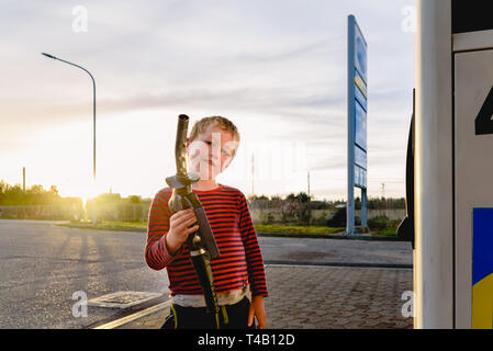 Bambino tenere il tubo di una pompa del carburante al tramonto in corrispondenza di una stazione di gas. Foto Stock