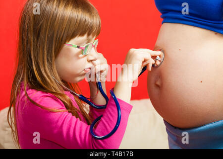 La gravidanza, il concetto di famiglia. Figlia utilizzando uno stetoscopio sulla sua madre gravida pancia per sentire i gemelli i toni del cuore Foto Stock