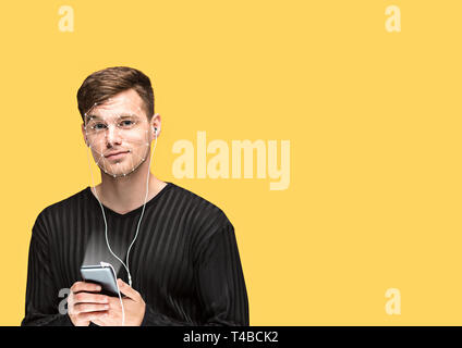 Aperto per il futuro. Un uomo con uno smartphone in camicia nera in piedi su sfondo giallo. La nuova tecnologia di riconoscimento facciale su griglia poligonale. Concetto di cyber security, business, lavoro. Copyspace. Foto Stock