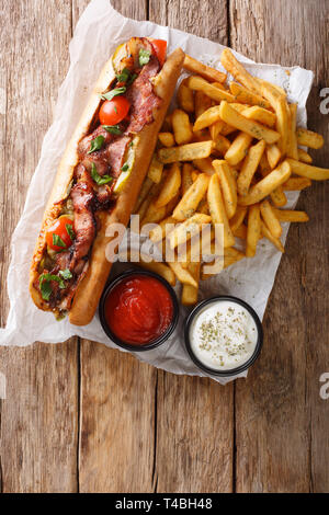 Stile rustico di hot dog con pancetta e verdure servita con patatine fritte e salse close-up sul tavolo. Verticale in alto vista da sopra Foto Stock