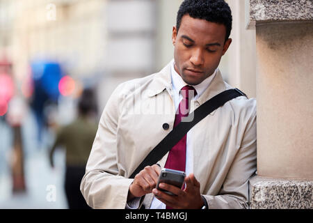 Giovane imprenditore nero appoggiato su di una parete in strada a Londra utilizzando lo smartphone, il fuoco selettivo close up Foto Stock