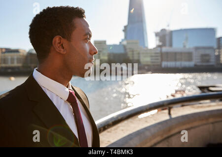 Giovane imprenditore nero che indossa camicia e cravatta in piedi dal fiume Thames, London, guardando lontano, retroilluminato Foto Stock