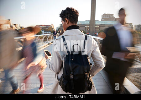 Vista posteriore del giovane adulto donna in piedi sul Millennium Bridge, Londra, utilizza lo smartphone, sfocatura del movimento Foto Stock