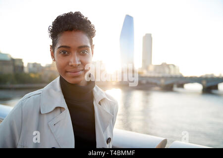 Giovane nero donna che indossa un maglione turtleneck e un mackintosh in piedi sul Millennium Bridge, Londra, cercando di fotocamera a sorridere Foto Stock