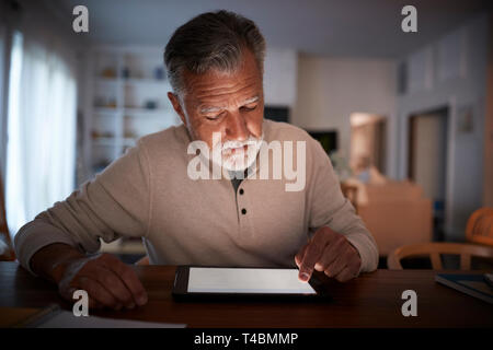 Senior uomo ispanico seduta a una tavola utilizzando un computer tablet a casa di sera, close up Foto Stock