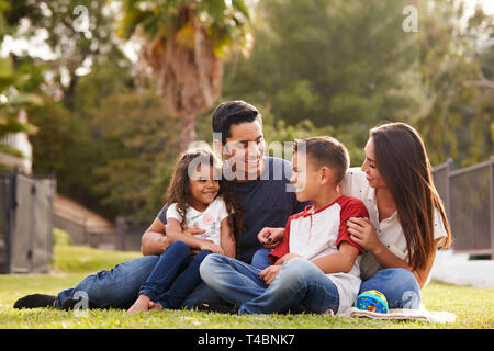 Felice giovane famiglia ispanica seduti insieme sull'erba nel parco, guardando ogni altro Foto Stock