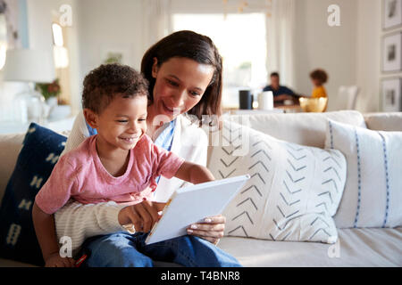 Close up della giovane madre seduta su un divano in soggiorno con il suo bambino sul suo ginocchio, lui la lettura di un libro, padre e figlia seduti ad un tavolo in background, la messa a fuoco su oggetti in primo piano Foto Stock