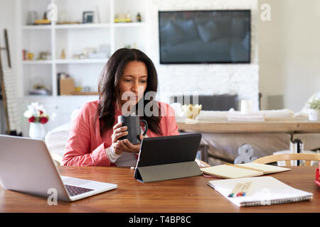 Donna di mezza età seduti a un tavolo di lettura con un computer tablet, tenendo in mano un bicchiere, vista frontale Foto Stock