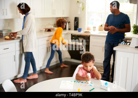 Il Toddler boy pittura seduti ad un tavolo in cucina pittura una foto, la sua famiglia occupato in background Foto Stock