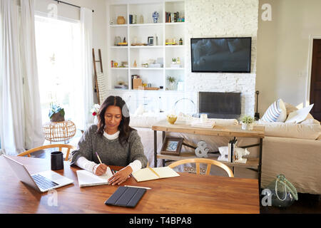 Felice di mezza età razza mista donna seduta al tavolo nella sua sala da pranzo rendendo note, vista in elevazione Foto Stock