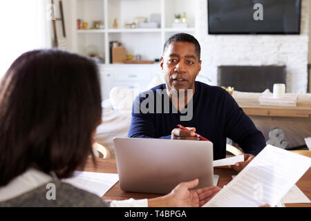 Uomo millenario con computer portatile dando consigli finanziari per una donna seduta al tavolo in possesso di un documento nella sua sala da pranzo, vicino il fuoco selettivo Foto Stock