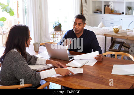 Uomo millenario con computer portatile dando consigli finanziari per una donna seduta al tavolo in possesso di un documento nella sua sala da pranzo, vista laterale Foto Stock