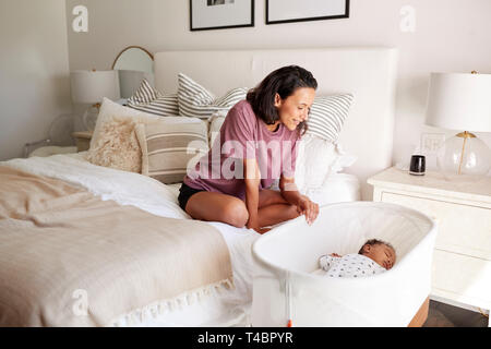 I giovani adulti madre seduta sul suo letto guardando verso di lei in tre mesi il bambino dorme nella sua culla Foto Stock