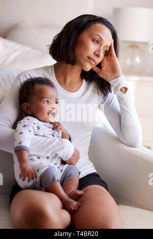 I giovani adulti madre seduto in poltrona nella sua camera da letto, tenendo le sue tre mesi figlio, guardando lontano nella contemplazione, verticale Foto Stock