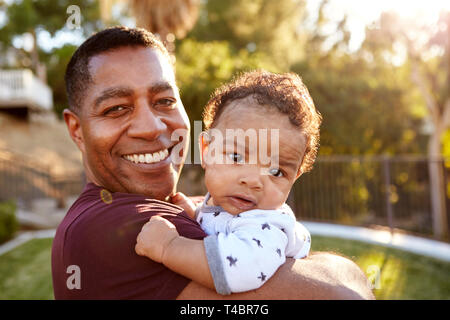 Fiera Millenaria di nonno in piedi nel giardino tenendo la sua tre mesi al nipote e sorridente alla telecamera, vicino, testa e spalle Foto Stock