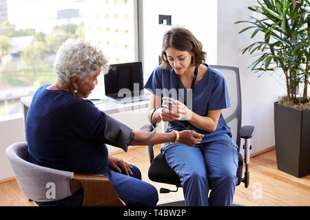 L'infermiera indossando Scrubs In ufficio controllo Senior le pazienti di sesso femminile della pressione del sangue Foto Stock