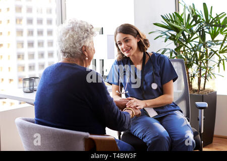 L'infermiera indossando Scrubs In ufficio rassicurante Senior paziente femmina e tenendo le Sue mani Foto Stock