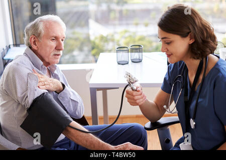 L'infermiera indossando Scrubs In ufficio controllo Senior i pazienti di sesso maschile della pressione del sangue Foto Stock