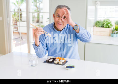 Bello il senior uomo mangiare take away sushi utilizzando bastoncini a casa con la faccia felice sorridente facendo segno OK con la mano sull'occhio guardando attraverso le dita Foto Stock