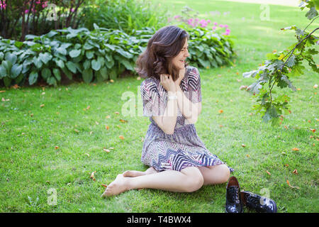 Giovane e bella brunete donna con capelli lunghi marrone in appoggio e sorridente sul parco o giardino sfondo verde. Donna europea. Ritratto di felice teenage Foto Stock