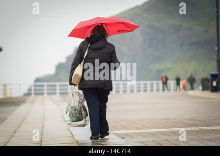 Aberystwyth Wales UK, martedì 16 aprile 2019. Regno Unito Meteo : Una donna camminare sotto un ombrello rosso lungo la passeggiata su una noiosa e umido ma mite giornata al mare in Aberystwyth Wales come la Pasqua vacanze scolastiche entrare in oscillazione. Credito foto Keith Morris / Alamy Live News Foto Stock