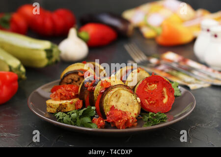 Ratatouille di verdura cotta: melanzane, zucchine e pomodori si trova su una piastra su un sfondo scuro, sul tavolo ci sono posate, sale, Foto Stock