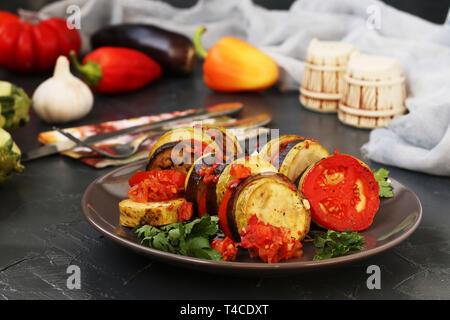 Ratatouille di verdura cotta: melanzane, zucchine e pomodori si trova su una piastra su un sfondo scuro, sul tavolo ci sono posate, sale, Foto Stock